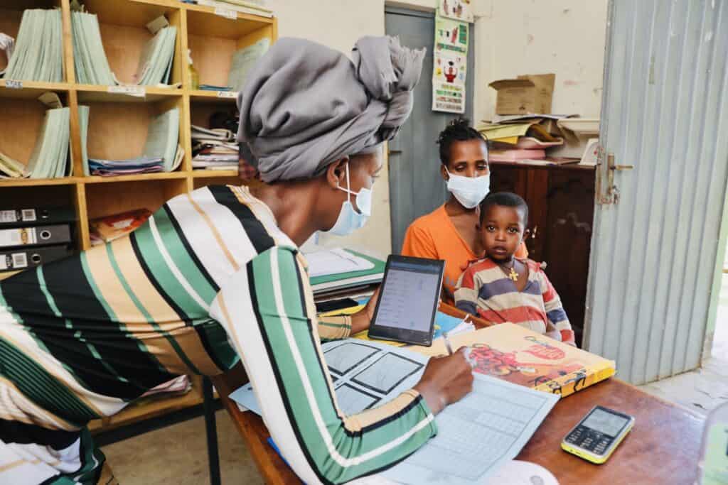 A female health worker refers to a tablet while attending to a woman and her child in an African medical office.