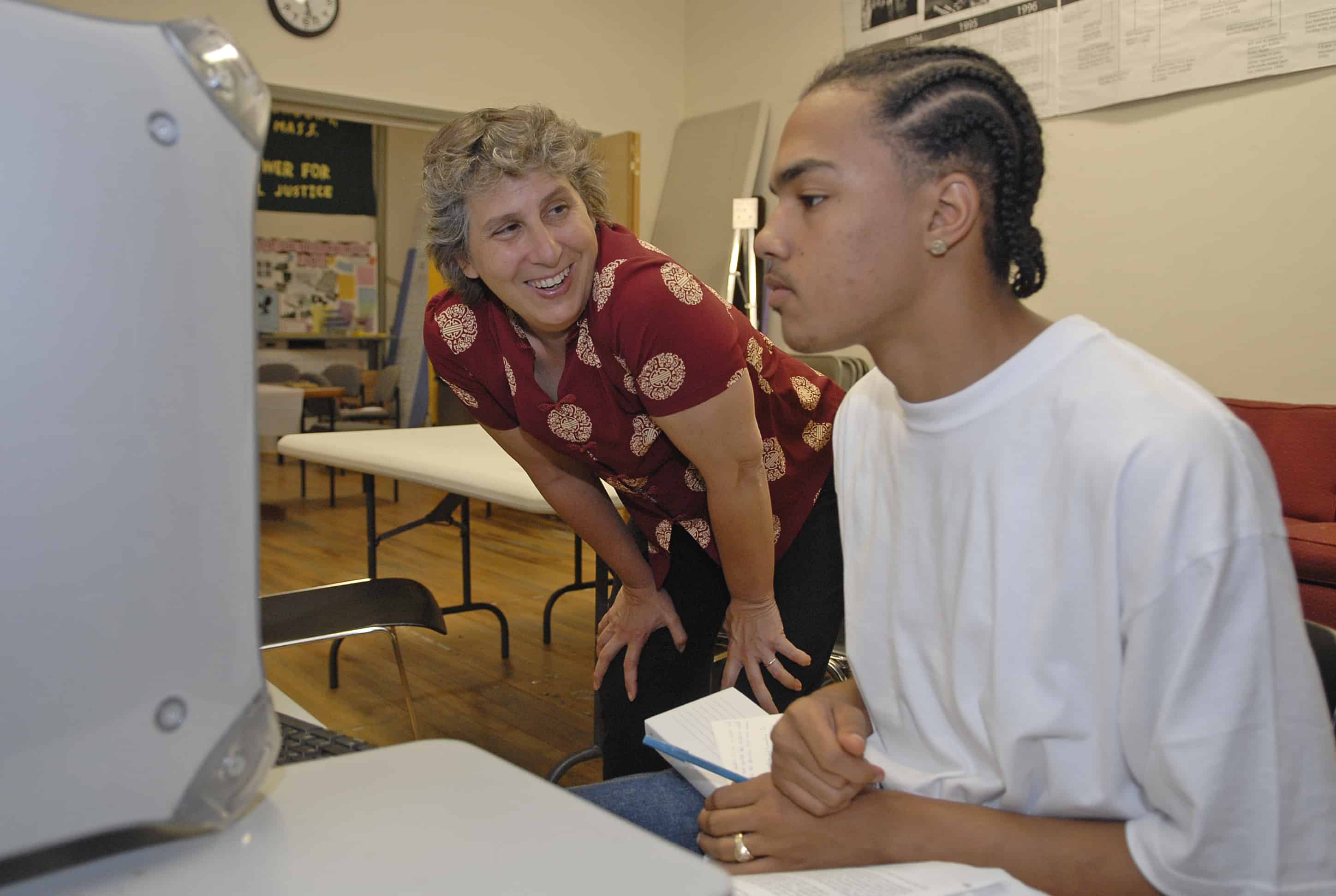 A middle-aged white woman wearing a red shirt leans down to talk to a student working on a computer.