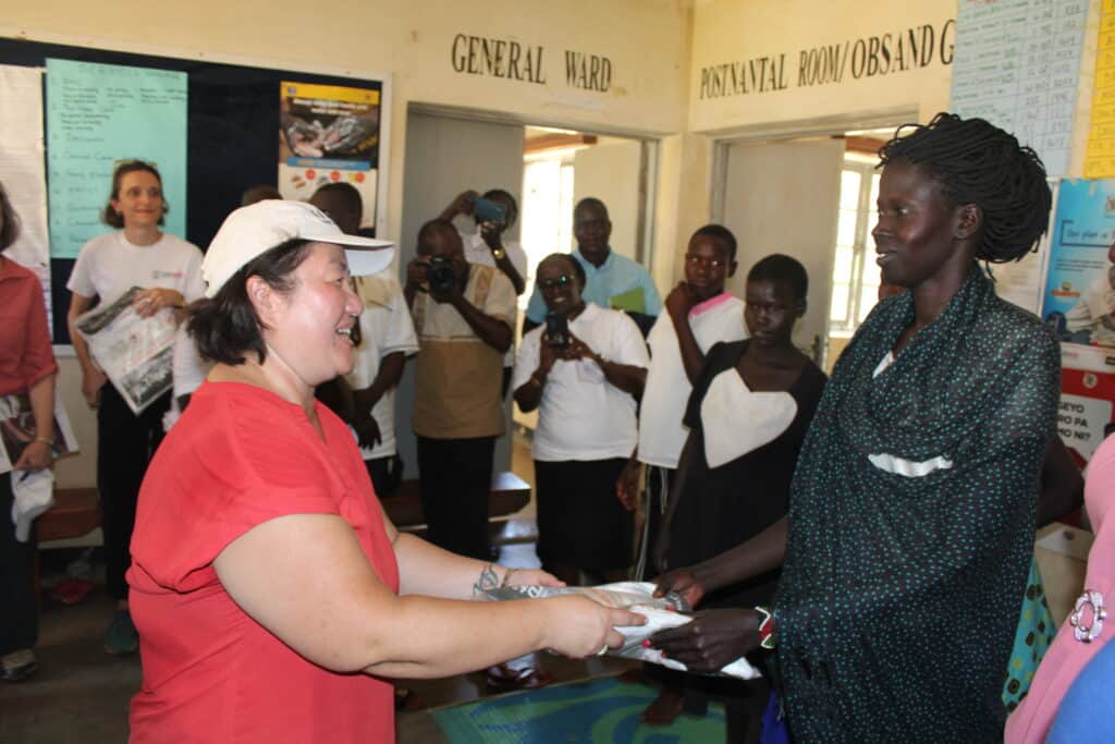 Mia Beers, USAID deputy assistant administrator in the Bureau for Resilience, Environment, and Food Security, hands a treated mosquito net to a pregnant woman at Palabek Ogili Health Centre III. Pregnant women are given mosquito nets so they can protect themselves and their unborn babies from mosquitos and the diseases they spread.