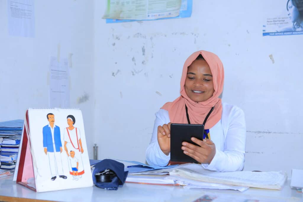 A female health worker sits at a table reviewing information on a tablet.