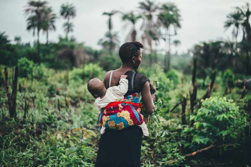 a woman carries a baby on her back in a rainforest setting.